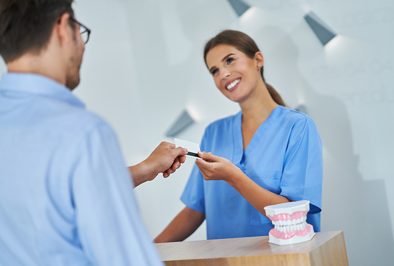 The image shows a professional setting where a dental receptionist is handing a card to a patient at a counter.